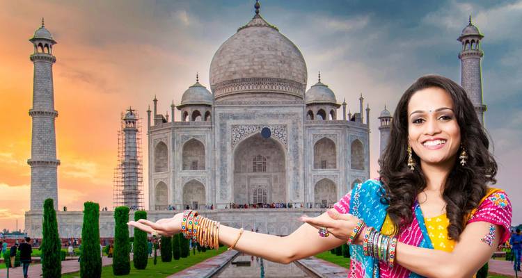 A woman in traditional attire smiling with the Taj Mahal in the background at sunset.