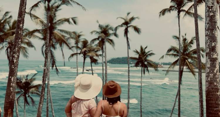 Two women looking out at the ocean with tall palm trees in the foreground.