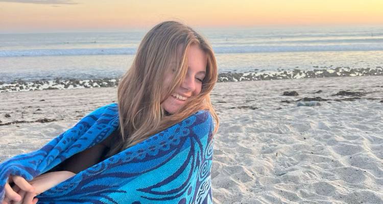 Woman smiling, wrapped in a blue towel on a sandy beach at sunset.