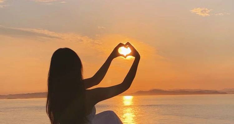 Woman making a heart shape with her hands at sunset over the sea.