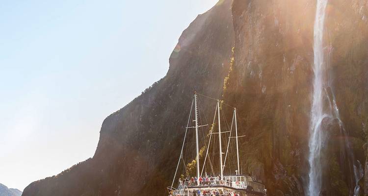 Un bateau touristique navigue sous une grande cascade qui se déverse des parois rocheuses abruptes dans le fjord de Milford Sound.