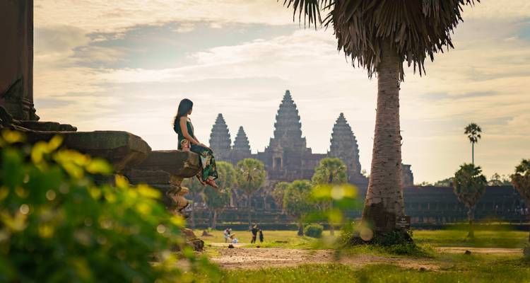 Femme assise près d'Angkor Vat avec des palmiers et une ambiance de lever de soleil.