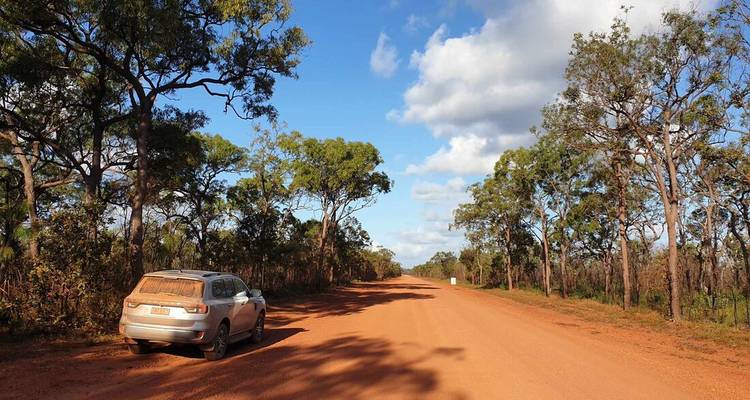 Chemin de terre rouge avec un véhicule sur le côté entouré d'arbres.