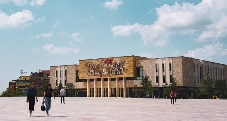 Großes Gebäude mit einem dekorativen Wandgemälde an einem zentralen Platz, Menschen gehen spazieren.