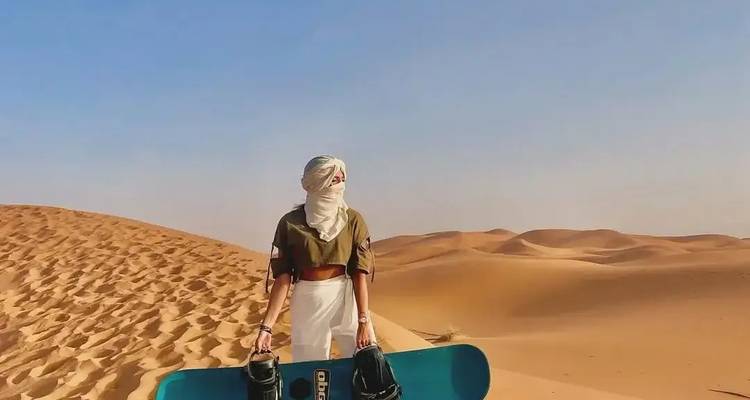 Person holding a snowboard, standing on sand dunes under a clear sky.