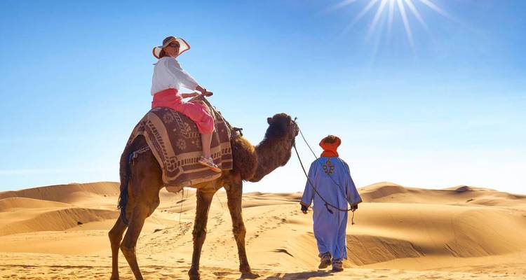 Person riding a camel guided by another person across sand dunes under the sun.