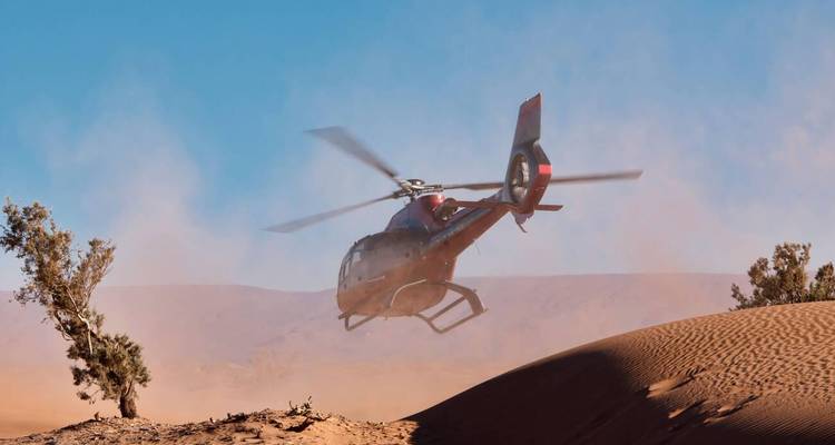A helicopter taking off in a desert area with sand dunes.