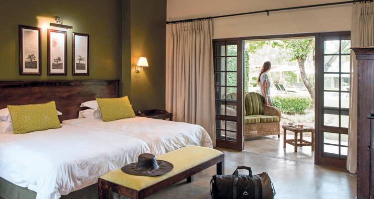 Cómoda habitación de lodge con dos camas individuales, muebles rústicos, y una mujer de pie en el patio mirando el jardín.