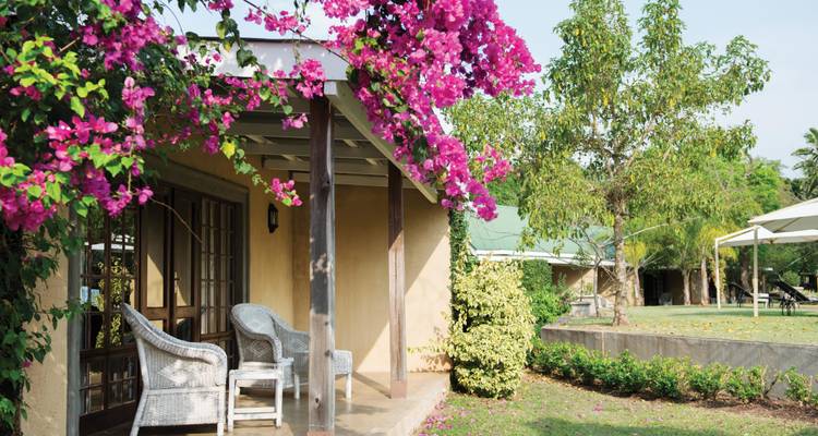 Veranda de la cabaña con sillas de mimbre cubiertas de buganvilia rosa vibrante que da a jardines bien cuidados.