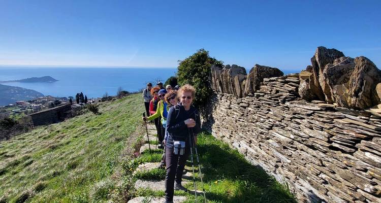 Eine Reihe von Wanderern lächelt neben einer Steinterassenmauer hoch über der ligurischen Küste unter blauem Himmel.