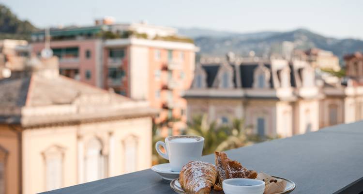 Cappuccino und blätterteigige Croissants ruhen auf einem Balkontisch mit Blick auf pastellfarbene ligurische Dächer.