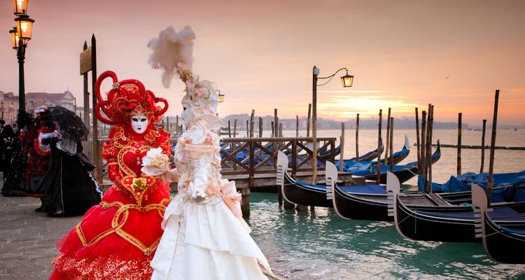 Masked figures in ornate costumes at a Venetian canal during sunset.