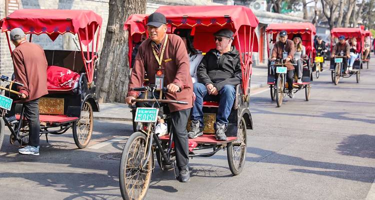 Fila de triciclos tradicionales pedaleando por un hutong histórico con turistas sentados bajo toldos rojos.