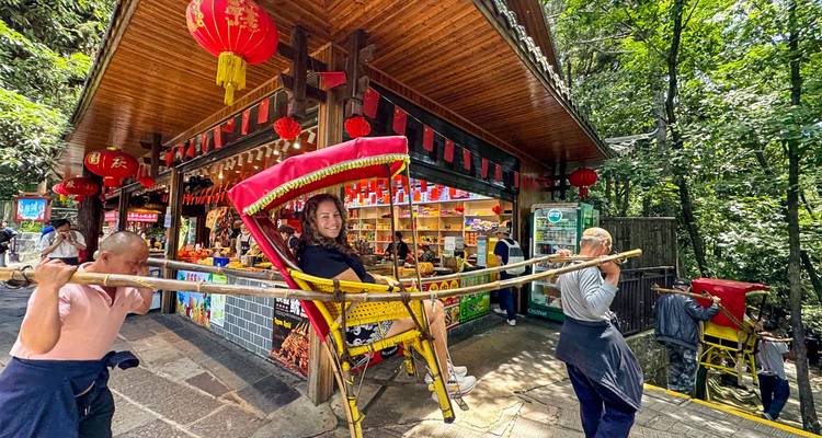El viajero disfruta de un paseo en silla de bambú cargada por porteadores junto a un puesto de recuerdos rodeado de bosque.