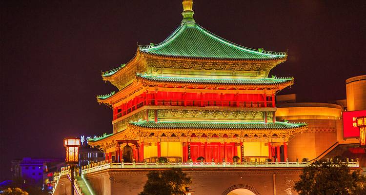 La Torre de la Campana de Xi'an brilla en tonos esmeralda y oro contra el cielo nocturno, iluminada por focos de luz vibrantes.