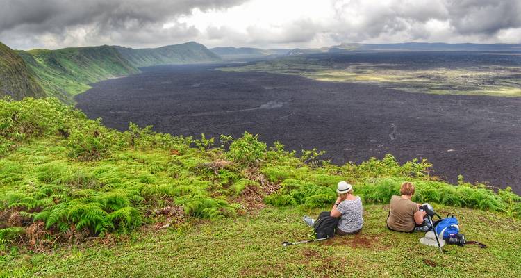 Two hikers rest on a lush green rim overlooking a vast black lava plain under dramatic clouds.