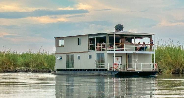 Houseboat on a river with reeds and a cloudy sky.