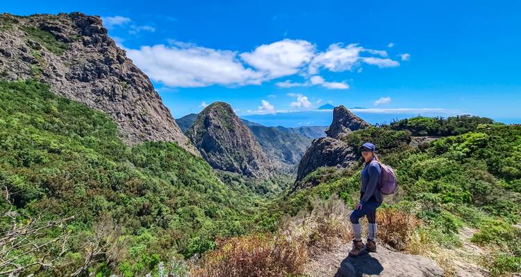 Un excursionista se para en una saliente rocosa con vistas a exuberantes valles volcánicos y picos dramáticos bajo un cielo azul vibrante.