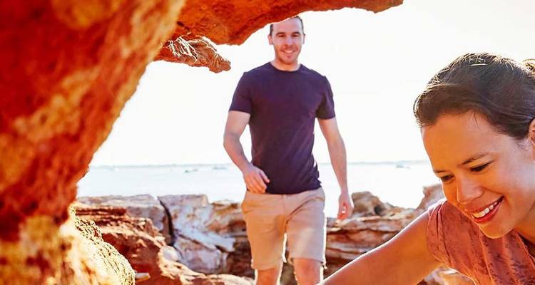 Couple exploring colourful layered rock formations near the coast under bright sunlight.
