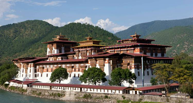 Das elegante Kloster Punakha Dzong erstreckt sich über den Fluss mit bewaldeten Hügeln und blauem Himmel dahinter.