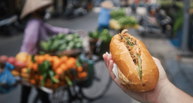 Un gros plan d'un sandwich banh mi avec un marché de rue animé en arrière-plan.