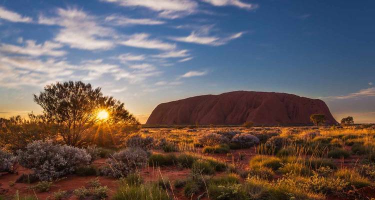 La luz del amanecer ilumina el monolito de Uluru y la maleza del desierto bajo nubes dispersas.