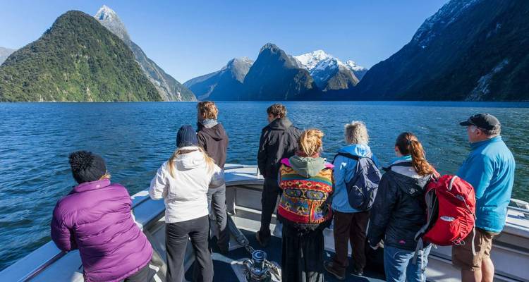Des voyageurs se tiennent sur le pont d'un bateau admirant les falaises du fjord et les sommets enneigés reflétés dans l'eau claire.