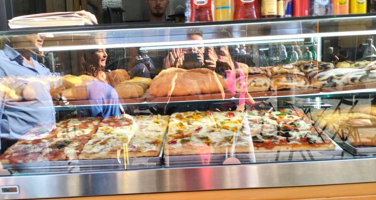 Vitrine de boulangerie avec différents types de pizzas et reflet de personnes.