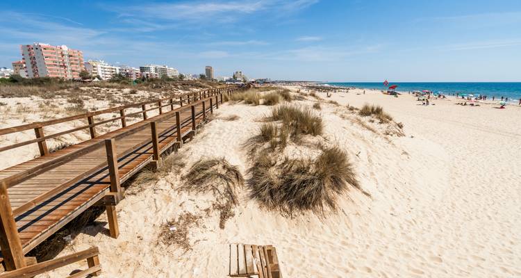 Une promenade en planches et une plage avec des baigneurs de soleil et des bâtiments au loin.