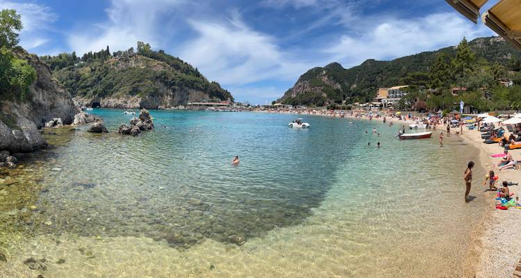 Überfüllter Strand mit klarem Wasser und umliegenden Hügeln.
