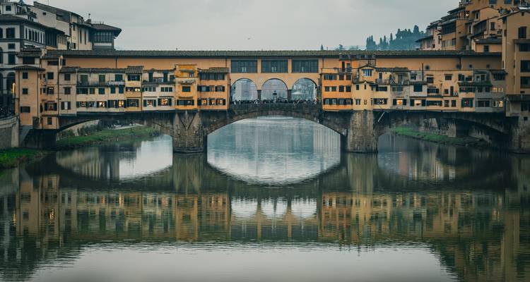 Ponte Vecchio reflecting on the Arno River under a cloudy sky