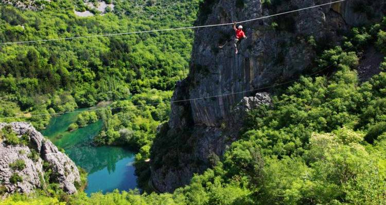 Person crossing a zipline over a lush green canyon with a river below.