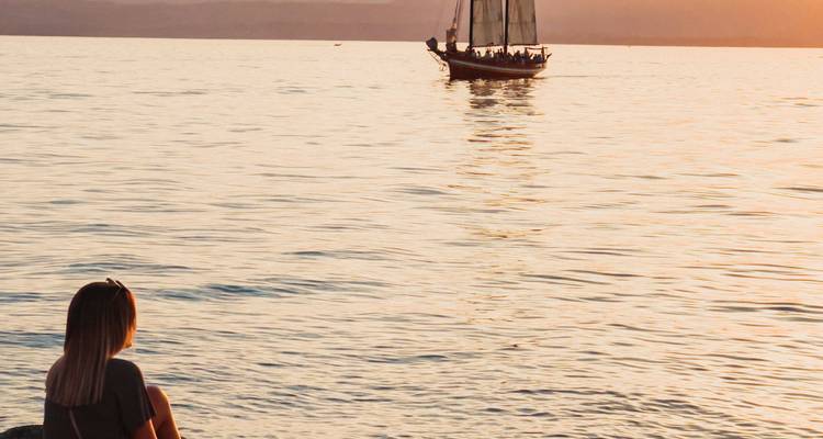Velero en un mar tranquilo durante el atardecer con una persona sentada junto al agua.