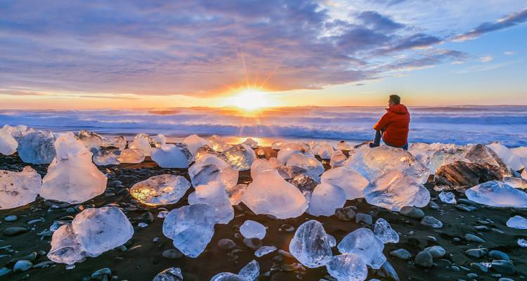 Person sitting among ice chunks on a black sand beach at sunset.