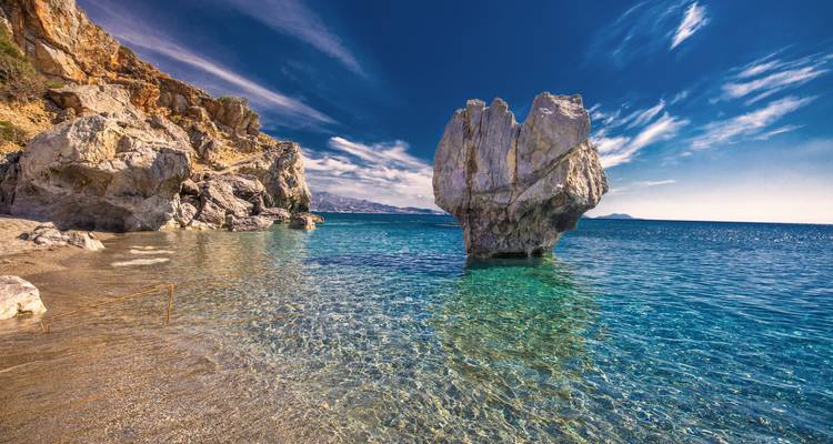 Rocky shore with a large stone formation in clear blue water.