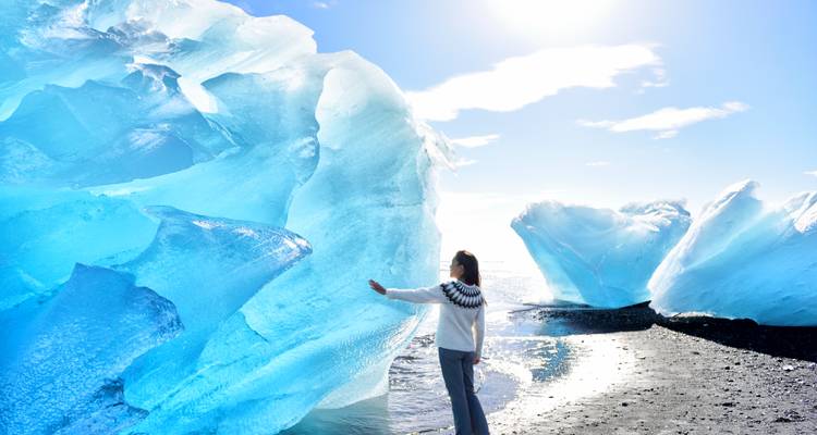 Person touching a large piece of blue ice near the ocean, with bright sunlight.
