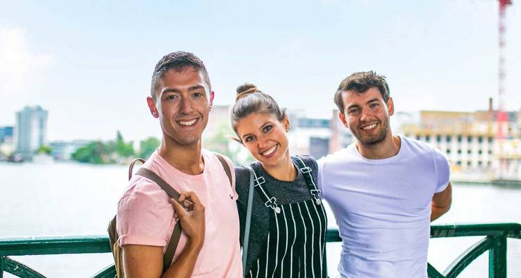 Drei Freunde stehen neben funkelndem Wasser mit moderner Amsterdamer Skyline im Hintergrund