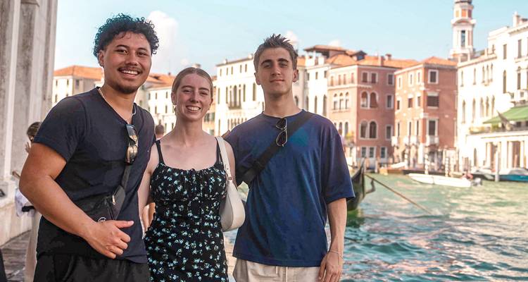 Tres amigos posan junto al agua esmeralda frente a elegantes edificios venecianos