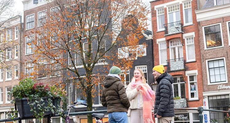 Three friends chat on a bridge beside canal houses and autumn foliage in Amsterdam.