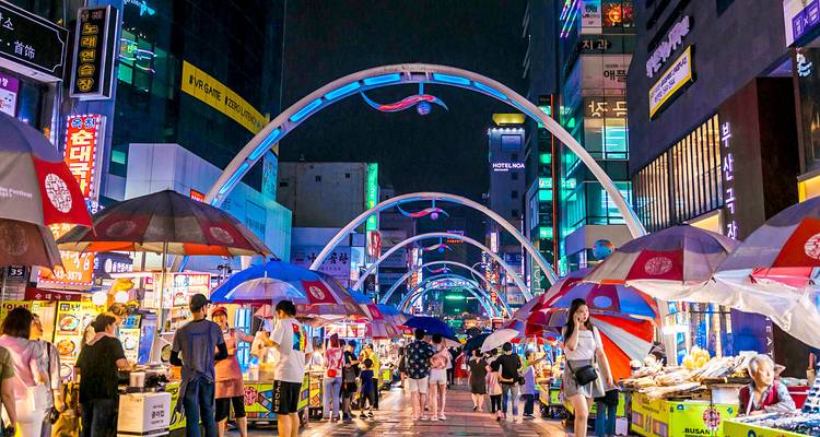 Scène de rue animée d'un marché nocturne remplie de monde et d'étals de nourriture.