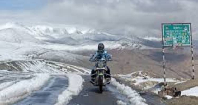 Motorcyclist on a snow-covered road with a sign and mountains in the background.