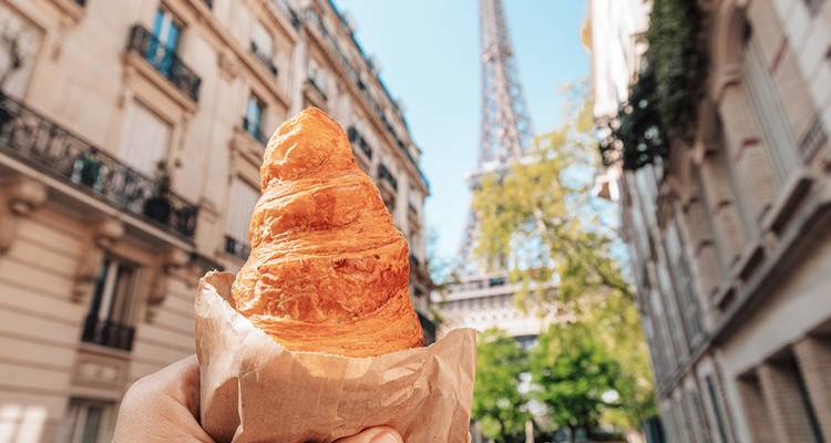 Nahaufnahme einer Hand, die ein Croissant hält, mit dem Eiffelturm sichtbar entlang einer Pariser Straße.
