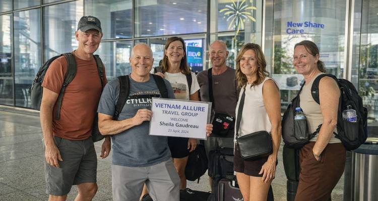 Sechs Reisende lächeln in einer Flughafen-Ankunftshalle und halten ein Willkommensschild für ihre Vietnam-Tour.