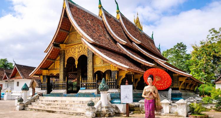 Frau in traditioneller laotischer Kleidung mit rotem Sonnenschirm posiert vor einem kunstvollen Tempel mit goldenem Dach.