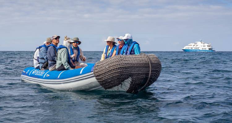 Group of people in a small boat on the ocean with a larger vessel in the background.