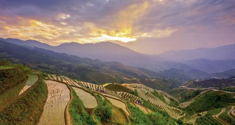 Terrassenförmige Reisfelder unter einem stimmungsvollen Sonnenuntergang mit Bergen im Hintergrund.