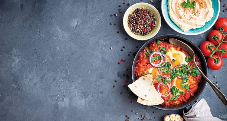 Flat lay of a bowl of shakshuka with pita bread and tomatoes, accompanied by spices and a cloth napkin.