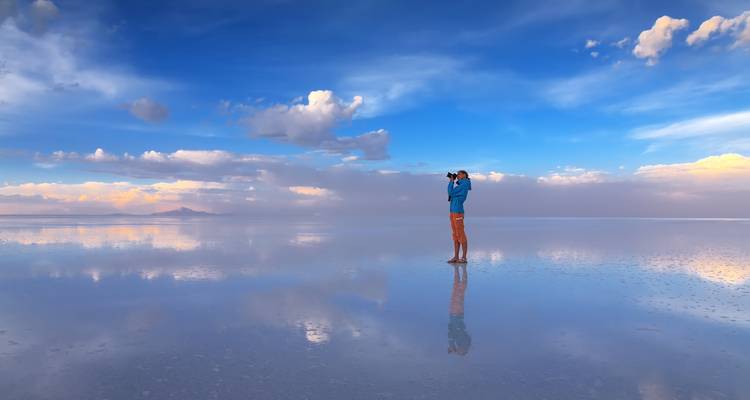 Een eenzame fotograaf staat op de glanzende spiegel van het Uyuni-zoutmeer onder een enorme blauwe hemel.