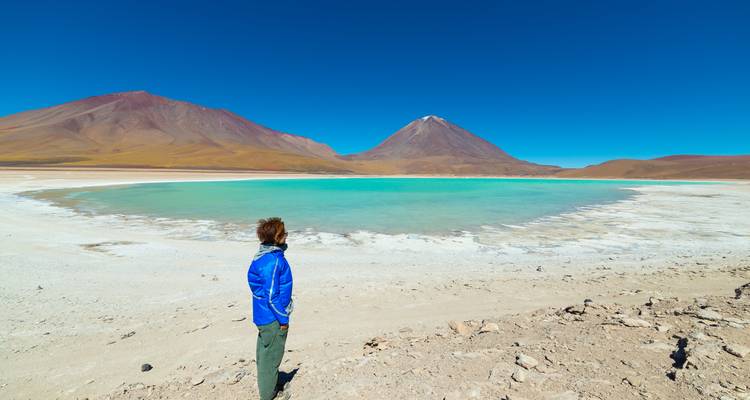 Un viajero con chaqueta azul admira las aguas verde esmeralda de la Laguna Verde respaldadas por un cono volcánico.