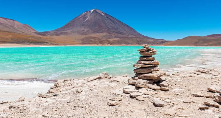 Un montículo de piedras apiladas en la orilla blanca se asoma a las aguas turquesas brillantes de la Laguna Verde con un volcán de fondo.